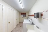 This shared laundry room is located within the residential community at 458–498 Sandra Ln in North Syracuse, New York. The space is thoughtfully laid out to maximize functionality within its compact footprint. The room features multiple top-loading washing machines and a dryer positioned along one wall, accompanied by a deep utility sink for added convenience. A wall-mounted paper towel dispenser adds a practical touch for residents.

The interior is finished with cream-colored walls accented by bold red and gray horizontal stripes, giving the space a lively, welcoming character. A playful wall decal reading "Laundry Room — 5¢ — Repeat" adds a charming, lighthearted element to the décor. Wire shelving brackets along the wall provide space for residents to organize laundry supplies and personal items.

Flooring consists of durable speckled terrazzo-style material, well-suited for a high-traffic utility area. A single window dressed with a red toile valance allows natural light to filter in, brightening the room. Overhead fluorescent lighting ensures the space remains well-illuminated at all times. A baseboard heater beneath the window offers warmth during colder months. The room is accessed through a distinctively brown door clearly labeled "Laundry Room," connecting conveniently to the broader building corridor.