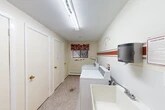 This shared laundry room is located within the residential community at 458–498 Sandra Ln in North Syracuse, New York. The space is thoughtfully laid out to maximize functionality within its compact footprint. The room features multiple top-loading washing machines and a dryer positioned along one wall, accompanied by a deep utility sink for added convenience. A wall-mounted paper towel dispenser adds a practical touch for residents.

The interior is finished with cream-colored walls accented by bold red and gray horizontal stripes, giving the space a lively, welcoming character. A playful wall decal reading "Laundry Room — 5¢ — Repeat" adds a charming, lighthearted element to the décor. Wire shelving brackets along the wall provide space for residents to organize laundry supplies and personal items.

Flooring consists of durable speckled terrazzo-style material, well-suited for a high-traffic utility area. A single window dressed with a red toile valance allows natural light to filter in, brightening the room. Overhead fluorescent lighting ensures the space remains well-illuminated at all times. A baseboard heater beneath the window offers warmth during colder months. The room is accessed through a distinctively brown door clearly labeled "Laundry Room," connecting conveniently to the broader building corridor.