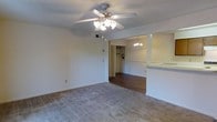 This single-floor space offers an open and functional layout that connects the living and dining areas seamlessly. The living area is finished with wall-to-wall carpet, providing a comfortable and warm underfoot feel, while a multi-blade ceiling fan with an integrated light fixture anchors the space from above and ensures adequate air circulation and illumination.

A transitional opening leads into the adjacent dining area, where the flooring shifts to rich hardwood, creating a natural visual distinction between the two zones. A chandelier hangs above the dining space, adding a touch of elegance. A half-wall or breakfast bar with a light-colored countertop is visible along one side, suggesting a connection to the kitchen and offering additional utility for casual dining or entertaining.

Beyond the dining area, a closet with bifold doors is visible, offering convenient built-in storage. The walls throughout are finished in a neutral white tone, contributing to a bright and clean atmosphere. The textured ceiling adds subtle depth to the interior. Overall, the space presents a practical and cohesive design that balances comfort with everyday functionality.
