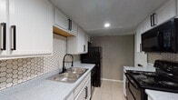 This two-story space is designed with a consistent, neutral aesthetic throughout, featuring warm gray walls, white trim, and light ceramic tile flooring that extends across the majority of the layout. The main level offers an open living area anchored by a sliding glass door dressed with vertical blinds, allowing natural light to filter in while providing access to an outdoor area with visible greenery. A carpeted staircase with a white railing connects the two floors, adding a transitional element to the layout.

The kitchen is a standout feature, outfitted with white cabinetry accented by matte black hardware, speckled quartz-style countertops, and a striking hexagonal tile backsplash. Black appliances, including a range, microwave, refrigerator, and dishwasher, provide a bold contrast to the bright cabinetry. A deep double-basin sink with a black pull-down faucet completes the functional workspace.

Additional rooms on the main level include generously sized spaces with tile flooring, ceiling fans, and ample natural light from windows and sliding doors. A bathroom features a tub-shower combination with white tile surround, tile flooring, a speckled vanity countertop, and matte black fixtures. A closet adjacent to the bathroom adds practical storage.
