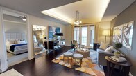 This thoughtfully designed single-floor living space combines contemporary elegance with functional comfort. The living area is anchored by rich, dark hardwood flooring that extends throughout, creating a warm and cohesive foundation. A defining architectural feature is the tray ceiling, which is enhanced by integrated accent lighting that adds depth and a soft ambient glow to the room.

Natural light fills the space through a set of French doors that lead to a private balcony, offering views of the surrounding exterior. Sheer roller shades provide flexible light control while maintaining an airy atmosphere. A stylish multi-bulb globe pendant light serves as a focal point overhead, complementing the overall modern aesthetic.

The living area is furnished with a mix of textured seating, including a striped accent chair and upholstered pieces arranged around a round tufted ottoman. A geometric, multi-toned area rug grounds the seating arrangement with vibrant warmth. A mirrored console and large floor mirror add visual dimension, while abstract wall art contributes a curated, gallery-like quality to the space. The overall ambiance is polished, inviting, and well-appointed.