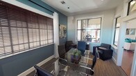 Located at 8401 Medlock Dr in Fort Worth, Texas, this single-floor professional office space combines modern design with functional work environments. The space is anchored by a well-appointed conference room featuring a sleek glass-top table surrounded by contemporary chrome-framed chairs. Bold teal accent walls contrast with crisp white trim and neutral tones, creating a polished yet approachable atmosphere. Large windows fitted with wooden blinds and sheer white curtains allow ample natural light to filter in, enhancing the overall brightness of the space. A distinctive interior window connects the conference room to an adjoining area, adding architectural interest and visual openness between rooms. Abstract wall art and a large statement clock with a Roman numeral iron frame contribute to the space's curated aesthetic. A dedicated workstation area provides multiple computer monitors, a granite-topped desk surface, and built-in storage, making it well-suited for administrative tasks. Wood-look flooring and patterned carpet complement the contemporary design scheme throughout. Additional seating, including leather accent chairs and a compact side cabinet, round out the furnishings, offering flexible arrangements for meetings or private consultations. The overall ambiance is professional, modern, and thoughtfully designed for a productive work environment.