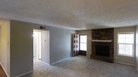 This single-story space presents a straightforward and functional layout centered around a spacious living area. The room is finished with neutral wall-to-wall carpeting that adds warmth underfoot, while the textured ceiling provides a classic architectural detail overhead. A ceiling-mounted HVAC vent ensures year-round climate control throughout the space.

The most prominent feature of the living area is a floor-to-ceiling brick fireplace positioned along one wall, serving as a natural focal point. The stacked brick surround extends to the ceiling and includes a mantel shelf, adding both visual interest and a sense of warmth to the room. Adjacent to the fireplace, a door with a decorative cutout panel provides access to an outdoor area, allowing natural light to filter into the space.

A doorway on the opposite side of the room leads to an adjacent area, which appears to include additional rooms and a hallway. The walls are painted in a soft, neutral gray tone that complements the earthy tones of the brick fireplace. The overall ambiance of the space is understated and practical, with key features that provide comfort and character in an uncluttered setting.