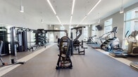 This contemporary fitness center is designed to support a full range of workout routines in a bright, professionally appointed environment. The space is housed on a single floor and features soaring ceilings that contribute to an open, energizing atmosphere. Linear LED lighting runs the length of the ceiling, supplemented by pendant fixtures, ensuring consistent, well-distributed illumination throughout the facility.

The layout is thoughtfully organized to separate functional zones. The strength training area is outfitted with a multi-station cable machine, a functional trainer, weight benches, stability balls, battle ropes, and a fully stocked dumbbell rack. Floor-to-ceiling mirrors line the walls, enhancing spatial perception and supporting proper form during workouts. The cardio zone features multiple treadmills, including Assault Fitness and Star Trac models, alongside stationary bikes and additional cardio equipment.

Durable rubber flooring covers the workout areas, providing both safety and comfort. Large windows along multiple walls flood the space with natural light and offer views of the surrounding exterior. Wall-mounted flat-screen televisions are positioned for visibility throughout the room. The neutral gray walls and clean finishes lend the space a polished, modern aesthetic befitting a high-quality fitness facility.