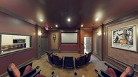This private theater is thoughtfully designed to deliver an authentic cinematic experience within a refined and intimate setting. The room is anchored by a large projection screen flanked by elegant burgundy curtains with decorative tassel tiebacks, creating a classic moviehouse aesthetic. The walls are finished in warm, deep red tones accented by detailed crown molding and wainscoting, lending the space a polished, upscale character. Recessed lighting in the dark ceiling provides carefully controlled illumination, supplemented by wall sconces that add a soft, ambient glow reminiscent of a traditional theater. Comfortable leather reclining chairs are arranged in a curved, tiered layout, ensuring excellent sightlines to the screen from every seat. Each chair features integrated cup holders, combining comfort with practicality. Decorative framed artwork adorns the walls between the doors, adding a cultivated touch to the overall design. Multiple entry doors are positioned on both sides of the room, providing convenient access. The flooring transitions from carpet in the main seating area to a decorative inlay near the screen, adding visual interest underfoot. Overall, the space balances functional theater design with warm, inviting décor for an exceptional viewing environment.