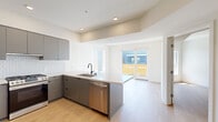 This contemporary single-floor residence showcases a clean, modern design characterized by crisp white walls, light wood flooring, and an abundance of natural light throughout. The open-concept kitchen is a standout feature, equipped with sleek flat-front cabinetry in a muted gray finish, stainless steel appliances including a full-size refrigerator and dishwasher, a gas range, and a peninsula with a white quartz countertop and an undermount sink with a matte black faucet. A geometric patterned tile backsplash adds a refined decorative touch to the cooking area. The kitchen flows seamlessly into the living space, which is bathed in natural light and opens directly onto a private balcony via full-height sliding glass doors, offering views of the surrounding urban landscape. The bedroom is generously proportioned, with large windows that frame scenic outdoor views and bring in ample sunlight. The bathroom is well-appointed with a soaking tub and tiled shower surround, a gray vanity with white countertop, a large mirror, and hardwood flooring that carries through from the rest of the residence. Additional features include in-unit laundry, recessed lighting, and a smart thermostat, completing this thoughtfully designed modern living space.