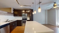 This well-designed single-floor residence offers a cohesive blend of modern finishes and functional living spaces. The kitchen serves as the centerpiece of the unit, showcasing rich dark-espresso cabinetry, white quartz countertops, and a classic white subway tile backsplash. A full suite of stainless steel appliances — including a refrigerator, electric range, and over-the-range microwave — complements the space, while a large kitchen island provides additional prep and seating space. Stylish pendant lighting hangs above the island, and adjustable track lighting illuminates the cooking area. Warm hardwood-style flooring flows throughout the kitchen and connecting areas, adding a sense of continuity. The bathroom is practical and clean in design, featuring a vanity with dark cabinetry and direct access to a dedicated walk-in closet with built-in shelving. Conveniently located nearby is an in-unit laundry area housing a full-size top-loading washer and dryer with additional wire shelving for storage. The bedroom is a quiet, neutral retreat finished with plush wall-to-wall carpeting and ample wall space, offering a comfortable and versatile environment. Throughout the unit, a neutral color palette and consistent finishes create a cohesive, modern atmosphere.