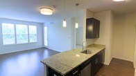 This single-floor residence offers a thoughtfully designed open-concept layout that seamlessly connects the kitchen and living areas, creating a spacious and inviting atmosphere. The kitchen is a standout feature, showcasing a large granite-topped island with a built-in stainless steel sink, elegant pendant lighting suspended from above, and rich dark cabinetry with sleek hardware that provides ample storage. The granite countertops display a classic light-toned pattern with subtle veining, adding a refined touch to the space. Warm hardwood flooring runs throughout, lending a cohesive and contemporary feel underfoot. Recessed and flush-mount ceiling lighting ensures the interior remains bright and well-illuminated. The living area benefits from natural light streaming in through multiple windows and a glass-paneled door, creating an airy, welcoming ambiance. Clean white walls provide a neutral canvas that complements the bold contrasts of the dark cabinetry and wood flooring. The open flow between the kitchen and living space makes this residence well-suited for both everyday living and entertaining, offering a modern yet comfortable environment throughout.