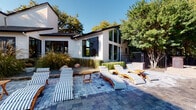 This single-story property blends modern farmhouse aesthetics with open, airy design throughout its thoughtfully arranged layout. The exterior showcases white horizontal siding with bold black-framed floor-to-ceiling windows, creating striking architectural contrast. A generously sized paver patio extends from the rear of the home, furnished with multiple teak-framed lounge chairs with striped cushions, ideal for outdoor relaxation. Native ornamental grasses and mature trees provide natural privacy and shade around the patio perimeter.

Inside, the entry foyer features shiplap-clad columns with sleek black-framed glass partitions that define spaces while maintaining visual openness. Light wood-look flooring runs consistently throughout the interior, complementing the neutral palette. The expansive main living area commands attention with dramatically vaulted ceilings and large windows that flood the space with natural light. Wide, low steps transition between interior levels, adding architectural interest.

The open-concept kitchen features white cabinetry, marble-look countertops, and a full suite of appliances, flowing seamlessly into the living area. A shiplap accent wall anchors the living space, where a wall-mounted television and statement pendant lighting complete the modern yet warm ambiance.
