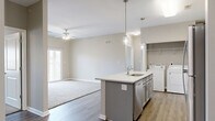 This single-floor, 3-bedroom, 2-bathroom residence offers a thoughtfully designed layout with modern finishes throughout. The open-concept kitchen and living area serves as the heart of the home, anchored by a central island with a granite-look countertop, undermount sink, and stylish pendant lighting. Grey shaker-style cabinetry pairs with a full suite of stainless steel appliances, including a refrigerator, electric range, and over-the-range microwave. Wood-look plank flooring extends throughout the kitchen and living areas, while plush carpeting adds warmth to the living room and all three bedrooms. The living room features a ceiling fan and access to a private outdoor balcony through sliding glass doors. A dedicated in-unit laundry area is conveniently tucked adjacent to the kitchen. The primary bedroom is generously sized and includes a ceiling fan and a walk-in closet. The two bathrooms are finished with grey vanity cabinetry, granite-look countertops, large mirrors, and earthy stone-look tile flooring. The additional bedrooms offer comfortable proportions with built-in closets. Neutral warm-grey walls throughout create a cohesive, move-in ready aesthetic that is both clean and inviting.
