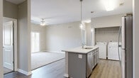 This single-floor, 3-bedroom, 2-bathroom residence offers a thoughtfully designed layout with modern finishes throughout. The open-concept kitchen and living area serves as the heart of the home, anchored by a central island with a granite-look countertop, undermount sink, and stylish pendant lighting. Grey shaker-style cabinetry pairs with a full suite of stainless steel appliances, including a refrigerator, electric range, and over-the-range microwave. Wood-look plank flooring extends throughout the kitchen and living areas, while plush carpeting adds warmth to the living room and all three bedrooms. The living room features a ceiling fan and access to a private outdoor balcony through sliding glass doors. A dedicated in-unit laundry area is conveniently tucked adjacent to the kitchen. The primary bedroom is generously sized and includes a ceiling fan and a walk-in closet. The two bathrooms are finished with grey vanity cabinetry, granite-look countertops, large mirrors, and earthy stone-look tile flooring. The additional bedrooms offer comfortable proportions with built-in closets. Neutral warm-grey walls throughout create a cohesive, move-in ready aesthetic that is both clean and inviting.