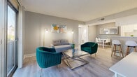 Located at 5175 Cinderlane Parkway in Orlando, Florida, this single-floor, 2-bedroom, 2-bathroom residence offers a thoughtfully designed layout that maximizes every square foot. Light wood-style flooring flows throughout, complementing a neutral gray color palette that creates a clean, cohesive aesthetic.

The living room is a bright, inviting space featuring a sliding glass door that opens to an outdoor balcony, flooding the room with natural light and providing outdoor views. A wall-mounted television and elegant gold-accented floor lamps round out the space.

Adjacent to the living area, the combined dining room and kitchen offer convenient everyday functionality. The dining area features a pass-through window connecting to the galley-style kitchen, which is outfitted with white cabinetry, laminate countertops, and full appliances including a range and refrigerator.

The primary bedroom is a comfortable retreat with a large window that brings in natural light and views of the surrounding greenery. The second bedroom is equally well-sized, finished in the same soft neutral tones with direct access to a bathroom.

Both bathrooms are finished in crisp white, each featuring a shower-tub combination. A dedicated closet provides additional storage, completing this efficiently laid-out Orlando residence.