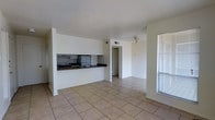 This single-floor space presents an open-concept layout that connects the kitchen and living areas in a practical, streamlined design. The kitchen is partially enclosed by a half-wall with a pass-through counter finished in dark laminate, providing separation while maintaining an open feel. The kitchen is equipped with a white refrigerator, a built-in stovetop with overhead ventilation, and a sink positioned along the counter. Light beige ceramic tile flooring runs consistently throughout the visible areas, contributing to a clean and cohesive aesthetic. The walls are painted in a neutral white throughout, keeping the space bright and versatile. A ceiling fan is mounted in the living area, offering both airflow and ambient lighting. A window fitted with horizontal blinds along one wall allows natural light to filter into the space. A doorway visible toward the rear of the unit leads to additional rooms, suggesting a thoughtful floor plan that balances communal and private areas. Overall, the space offers a functional, no-frills environment suited for everyday living.