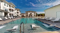 Located in Jacksonville, FL, this outdoor pool area offers a resort-inspired retreat designed for relaxation and recreation. The centerpiece is a generously sized swimming pool with crystal-clear blue water, complemented by built-in in-water lounge chairs that invite residents to unwind in true resort fashion. The pool is surrounded by a broad, well-appointed paver deck finished in a classic stone pattern, providing ample space for lounging and socializing. Rows of chaise lounge chairs and patio umbrellas line the poolside, offering shaded and sunny options throughout the day. A dedicated outdoor seating area with a dining table and chairs adds to the functional layout of the deck. Adjacent to the pool, a covered porch area features stylish wicker seating with plush cushions and accent pillows, accessible via large glass doors that connect to an interior space. The porch is finished with white shiplap siding, a painted ceiling, and classic wall-mounted lantern lighting, reflecting a coastal-inspired aesthetic. The pool area is enclosed by a black iron fence and is framed by multi-story residential buildings with balconies on all sides, creating a private, community-centered atmosphere beneath the expansive Florida sky.
