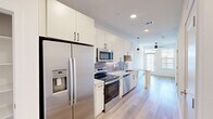 Located in Manor, TX, this thoughtfully designed one-bedroom, two-bathroom unit offers a modern aesthetic with efficient use of space across a single floor. The galley-style kitchen is a standout feature, outfitted with stainless steel appliances including a French door refrigerator with ice maker, electric range, built-in microwave, and dishwasher. White shaker cabinetry extends to the ceiling for ample storage, while granite countertops and a grey tile backsplash add a polished touch. Light wood-look flooring runs throughout the main living and kitchen areas, complemented by recessed lighting and pendant fixtures. The living area is brightened by natural light from a front-facing window and features a ceiling fan. A distinctive barn door connects the living space to the rest of the unit, adding a modern-rustic design element. The bedroom offers plush carpeting, a ceiling fan, and a window with blinds. A generously sized walk-in closet with built-in shelving and hanging rods provides excellent storage. The bathrooms are well-appointed, one featuring a soaking tub with tile surround, and the other offering a granite-topped vanity with white cabinetry and a large framed mirror.