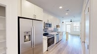 Located in Manor, TX, this thoughtfully designed one-bedroom, two-bathroom unit offers a modern aesthetic with efficient use of space across a single floor. The galley-style kitchen is a standout feature, outfitted with stainless steel appliances including a French door refrigerator with ice maker, electric range, built-in microwave, and dishwasher. White shaker cabinetry extends to the ceiling for ample storage, while granite countertops and a grey tile backsplash add a polished touch. Light wood-look flooring runs throughout the main living and kitchen areas, complemented by recessed lighting and pendant fixtures. The living area is brightened by natural light from a front-facing window and features a ceiling fan. A distinctive barn door connects the living space to the rest of the unit, adding a modern-rustic design element. The bedroom offers plush carpeting, a ceiling fan, and a window with blinds. A generously sized walk-in closet with built-in shelving and hanging rods provides excellent storage. The bathrooms are well-appointed, one featuring a soaking tub with tile surround, and the other offering a granite-topped vanity with white cabinetry and a large framed mirror.