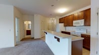 This single-floor residence is designed with an open layout that connects the kitchen and living area seamlessly. The kitchen is equipped with warm medium-toned wood cabinetry that extends to the upper and lower sections of the walls, providing ample storage space. White appliances, including a refrigerator, range, and over-the-range microwave, complement the light-colored countertops. A peninsula with a breakfast bar extends into the living space, featuring bar-height seating supports—ideal for casual dining or entertaining. The flooring transitions from a hard surface near the kitchen to plush carpeting in the living area, adding warmth underfoot. Recessed and flush-mount lighting fixtures illuminate the space evenly throughout. A doorway visible in the background leads to what appears to be a bathroom, showcasing matching wood cabinetry and vanity lighting. An additional door in the background, accented by a pendant light above, likely leads to a closet or entry point. The overall design is practical and cohesive, with neutral tones creating a calm and inviting atmosphere throughout.