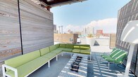 This rooftop terrace is a well-appointed outdoor space designed for urban living and entertaining, positioned high enough to command impressive panoramic views of a bustling downtown skyline. The terrace is divided into several distinct functional zones, each thoughtfully designed for a different outdoor experience.

The outdoor kitchen area features a stainless steel built-in gas grill set into a clean, white-plastered counter with an under-counter storage cabinet. Adjacent to the grill, a bar-height counter with sleek black stools provides seating for casual dining and socializing against the backdrop of surrounding city buildings.

A dedicated lounge area offers multiple reclining sun chairs arranged on a patch of artificial turf, framed by a large white privacy wall that doubles as a projection surface. Sculptural white planters add a modern decorative touch to this sun-drenched zone.

A covered seating nook, partially shaded by a pergola-style overhead structure and accented with weathered wood panel walls, houses a generous L-shaped sectional sofa with green cushions centered around a linear fire pit table. Drought-tolerant plantings, including agave, add a desert-appropriate landscape element throughout the terrace.