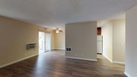 This single-floor residence offers a practical and well-appointed layout across several distinct living spaces. The open living and dining area is a standout, featuring dark hardwood-style flooring, warm beige walls, and a textured ceiling. A ceiling fan with a light fixture anchors the room, while a wall-mounted air conditioning unit provides climate control. Sliding glass doors lead to an outdoor area, allowing natural light to fill the space. A floor vent and additional window further enhance airflow and brightness.

The galley-style kitchen is efficiently designed with rich dark wood cabinetry lining both sides, granite-look countertops, a double stainless steel sink, and stainless steel appliances including a refrigerator, electric range, and over-the-range microwave. Light wood flooring and overhead lighting complete the functional workspace.

The bedrooms are carpeted for comfort, featuring neutral tones, baseboard heating, sliding closet doors, and windows that offer views of surrounding greenery. The bathroom includes a dark wood vanity with granite countertops, a large mirror with overhead lighting, a tiled tub-shower combination, and an adjacent walk-in closet area, offering a practical and organized layout.