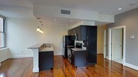 This single-floor space showcases a thoughtfully designed open-concept layout that seamlessly blends style and functionality. The kitchen is a standout feature, outfitted with rich, dark espresso cabinetry that extends to the ceiling, offering generous storage capacity. Granite countertops provide durable and attractive work surfaces, complemented by a full suite of stainless steel appliances including a refrigerator, electric range, and built-in microwave. A peninsula with a granite countertop and an integrated sink extends into the living area, creating a natural divide between the kitchen and the open living space while offering additional seating and prep area. Pendant lighting hangs above the peninsula, adding warmth and a modern touch to the kitchen zone. Recessed lighting throughout ensures the space is well-lit. The rich, warm-toned hardwood flooring flows continuously across the entire space, adding character and a sense of cohesion. The open layout and neutral wall tones create a bright, airy ambiance, while the clean architectural lines and modern finishes give the space a contemporary, polished feel overall.