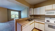 This single-floor residence offers a functional and comfortable layout designed for everyday living. The open-concept kitchen is equipped with white cabinetry featuring wood-tone trim, laminate countertops, a full suite of white appliances including an electric range, microwave, refrigerator, and dishwasher, and tile flooring. A peninsula counter connects the kitchen to the adjacent living area, creating a seamless flow between spaces. The spacious living room features plush carpeting, vaulted ceilings, a ceiling fan, and direct access to a private balcony through sliding glass doors, offering pleasant views of surrounding trees and greenery. A secondary window provides additional natural light. The primary bedroom is generously sized with soft carpeting, a walk-in closet, and an en-suite bathroom. A second bedroom also features carpet flooring and a large window overlooking the neighborhood. The bathroom includes a full tub-shower combination with white tile surround and tile flooring. An in-unit laundry area accommodates a full-size washer and dryer side by side, adding everyday convenience. Throughout the home, warm neutral tones on the walls complement the carpeted floors and create a cohesive, inviting ambiance.