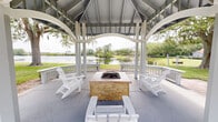 Located at 10010 Skinner Lake Dr in Jacksonville, Florida, this charming outdoor gazebo is a standout community gathering space designed for relaxation and socializing. The structure features a classic octagonal layout with a soaring vaulted roof supported by white-painted columns and defined by graceful arched openings on all sides, creating an open, airy atmosphere while providing shelter overhead.

At the center of the gazebo sits a substantial stone-clad fire pit table, serving as the focal point of the space. White Adirondack chairs are arranged around it, offering comfortable seating for residents and guests. The combination of the fire pit and surrounding seating makes this an inviting spot for both daytime gatherings and evening relaxation.

The gazebo opens to well-manicured green lawns, lush mature trees, and manicured landscaping. A standout feature is the direct view of a tranquil lake visible from within the structure, providing a peaceful natural backdrop that enhances the overall ambiance. The surrounding residential buildings, finished in classic gray siding, frame the scenery and contribute to the cohesive neighborhood aesthetic.

The concrete flooring is clean and well-maintained, and the white-painted woodwork throughout gives the space a bright, crisp, and welcoming character. This gazebo serves as an ideal outdoor retreat, blending architectural charm with natural surroundings.