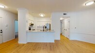 This well-appointed single-floor residence offers a clean, modern aesthetic defined by crisp white walls, light hardwood flooring, and thoughtful design details throughout. The open-concept layout connects the kitchen and living area seamlessly, creating a spacious and airy atmosphere ideal for both everyday living and entertaining. The kitchen is a standout feature, equipped with stainless steel Whirlpool appliances, including a range, over-the-range microwave, and refrigerator with ice dispenser. White shaker-style cabinetry with matte black hardware, a classic subway tile backsplash, and light stone countertops complete the polished look. A peninsula with a built-in sink provides additional prep space and casual seating. Crown molding accents the living area, adding an elegant architectural touch. The bedrooms are bright and comfortable, each featuring hardwood flooring and windows that invite natural light. The bathroom is finished in a consistent white palette, featuring a tub-shower combination with tile surround, a white vanity with stone countertop, and matte black hardware that ties the design together. The overall layout, as seen in the dollhouse view, is well-organized and functional, offering a cohesive living experience throughout.