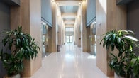 This single-floor lobby and amenity space showcases a thoughtfully designed contemporary interior that balances elegance with functionality. The grand entrance corridor immediately commands attention with its soaring double-height ceilings, warm light wood paneling on structural columns, and gleaming large-format white tile flooring that reflects abundant natural light streaming in through floor-to-ceiling glass entry doors at the far end.

Flanking the central corridor are intimate lounge alcoves, each distinctively furnished with bold, curated seating — including a rich terracotta sofa with patterned cushions and a vibrant mustard curved sofa paired with a woven leather accent chair — set against expansive windows that frame views of the surrounding urban landscape. These seating nooks are softly carpeted, providing a warm contrast to the polished tile of the main hall.

Lush tropical plants are strategically placed throughout, adding vitality and softness to the refined material palette of wood, tile, and muted grey tones. A dedicated fitness center is accessible directly from the lobby. An interior lounge area with rustic wood bench seating opens onto a landscaped outdoor terrace, complete with dining furniture, greenery, and open-sky views, offering a seamless indoor-outdoor connection ideal for relaxation.