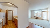 This single-floor residence offers a practical and comfortable layout thoughtfully designed to maximize functionality. The living area is bright and inviting, featuring plush wall-to-wall carpeting, a large window with an exterior door that floods the space with natural light, and a wall-mounted HVAC unit for year-round climate control. Warm hardwood-style flooring extends through the main corridor and into the kitchen area, providing a cohesive visual flow throughout.

The kitchen is accessed through a distinctive arched passageway, lending architectural character to the space. It features a galley-style configuration with warm wood cabinetry, light countertops, a double stainless steel sink, and a full suite of white appliances including a refrigerator, electric range, built-in microwave, and dishwasher. A dedicated bedroom offers carpeted flooring, a baseboard heater, and a window providing natural light and exterior views.

The full bathroom is equipped with a single-sink vanity, tile backsplash, a large wall mirror with overhead lighting, and a shower-tub combination. A separate in-unit laundry area accommodates a full-size washer and dryer. An additional utility room provides wire shelving for storage alongside the water heater, offering practical organizational space.