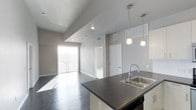 This single-floor space is designed with a clean, modern aesthetic that emphasizes openness and functionality. The open-concept layout seamlessly connects the kitchen and living area, creating a spacious, flowing environment ideal for both everyday living and entertaining.

The kitchen features a dark countertop island with a stainless steel double sink and a contemporary gooseneck faucet. Overhead, elegant pendant lights hang above the island, providing focused task lighting while adding a stylish touch. Built-in appliances are integrated beneath the counter, maintaining a streamlined look.

Dark hardwood flooring runs throughout the space, offering a bold contrast to the light gray walls and white trim. The smooth, polished floors contribute to the unit's contemporary feel while adding warmth and depth.

The living area is anchored by a large sliding glass door that invites an abundance of natural light, illuminating the open floor plan and creating a bright, airy ambiance. Recessed ceiling lighting complements the natural light throughout the day.

Additional features include a raised ceiling section in the living area that adds architectural interest and a sense of vertical space. Interior doors lead to separate rooms, providing well-defined private spaces within the otherwise open layout. The overall design is minimal, modern, and thoughtfully composed.