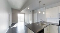 This single-floor space is designed with a clean, modern aesthetic that emphasizes openness and functionality. The open-concept layout seamlessly connects the kitchen and living area, creating a spacious, flowing environment ideal for both everyday living and entertaining.

The kitchen features a dark countertop island with a stainless steel double sink and a contemporary gooseneck faucet. Overhead, elegant pendant lights hang above the island, providing focused task lighting while adding a stylish touch. Built-in appliances are integrated beneath the counter, maintaining a streamlined look.

Dark hardwood flooring runs throughout the space, offering a bold contrast to the light gray walls and white trim. The smooth, polished floors contribute to the unit's contemporary feel while adding warmth and depth.

The living area is anchored by a large sliding glass door that invites an abundance of natural light, illuminating the open floor plan and creating a bright, airy ambiance. Recessed ceiling lighting complements the natural light throughout the day.

Additional features include a raised ceiling section in the living area that adds architectural interest and a sense of vertical space. Interior doors lead to separate rooms, providing well-defined private spaces within the otherwise open layout. The overall design is minimal, modern, and thoughtfully composed.
