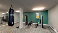 This fitness center is organized across a single floor and divided into several functional zones, providing a comprehensive workout environment. The main cardio area features a row of treadmills and elliptical trainers arranged along one side of the room, offering ample space for users to exercise comfortably. Patterned carpet tiles in neutral tones with teal accents cover the floors throughout, contributing to a cohesive and modern aesthetic.

A dedicated strength-training section houses a multi-station cable machine, a weight bench, and a resistance apparatus, while a hanging Everlast heavy bag adds a boxing element to the space. A dumbbell rack with an adjacent wall-mounted mirror provides additional free-weight options. The mirrored walls throughout the facility enhance the sense of openness and allow users to monitor their form.

A separate room, accessible through a wood-framed glass door, serves as a stretching and functional fitness area. It is equipped with stability balls, medicine balls, resistance bands, foam rollers, and step platforms. Instructional fitness posters are displayed on the teal accent walls. Flush-mounted fluorescent lighting illuminates the entire space evenly. A vending machine stocked with beverages is also available for convenience.