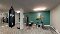 This fitness center is organized across a single floor and divided into several functional zones, providing a comprehensive workout environment. The main cardio area features a row of treadmills and elliptical trainers arranged along one side of the room, offering ample space for users to exercise comfortably. Patterned carpet tiles in neutral tones with teal accents cover the floors throughout, contributing to a cohesive and modern aesthetic.

A dedicated strength-training section houses a multi-station cable machine, a weight bench, and a resistance apparatus, while a hanging Everlast heavy bag adds a boxing element to the space. A dumbbell rack with an adjacent wall-mounted mirror provides additional free-weight options. The mirrored walls throughout the facility enhance the sense of openness and allow users to monitor their form.

A separate room, accessible through a wood-framed glass door, serves as a stretching and functional fitness area. It is equipped with stability balls, medicine balls, resistance bands, foam rollers, and step platforms. Instructional fitness posters are displayed on the teal accent walls. Flush-mounted fluorescent lighting illuminates the entire space evenly. A vending machine stocked with beverages is also available for convenience.
