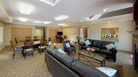 This single-floor community room is thoughtfully designed to serve as a comfortable and functional gathering space. The main lounge area makes a strong impression with a classic fireplace framed by a decorative mantel set against a rich, deep red accent wall. French doors nearby allow natural light to filter in, complementing the warm, inviting atmosphere. Plush leather sofas and accent chairs are arranged around glass-top coffee tables, creating an ideal setting for relaxed conversation and socializing.

The space opens into a generously sized dining area, where multiple wood dining tables and chairs are arranged to accommodate group gatherings and events. Patterned carpeting runs throughout the lounge and dining zones, adding warmth underfoot, while recessed and flush-mount ceiling lighting ensures the entire area is well-lit. Framed artwork adorns the walls, and blinds on the windows offer both privacy and light control.

Adjacent to the main gathering area is a functional kitchenette, finished with light oak cabinetry, a double stainless steel sink, and ample counter space. A coffee station and small appliances make it convenient for catering to events. Tile flooring in the kitchenette provides easy maintenance, and a paper towel dispenser and under-sink water filtration system reflect practical, everyday utility. Overall, the space balances comfort and functionality for community use.