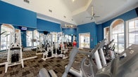This fitness center offers a spacious and energizing environment designed to support a full range of workout routines. The interior is defined by striking cobalt blue walls that contrast with crisp white crown molding and high vaulted ceilings, creating an open, airy atmosphere. Ceiling fans are strategically positioned throughout to maintain airflow during workouts.

The cardio zone is well-appointed with treadmills, elliptical machines, and a stationary bike, all positioned in front of large arched windows that flood the space with natural light and provide views of the outdoor surroundings. Wall-mounted televisions are conveniently placed throughout the room for entertainment during exercise.

The strength training area features a multi-station cable machine system offering a wide variety of resistance exercises. A dedicated free weights section includes a full dumbbell rack alongside stability balls in multiple sizes, catering to functional fitness and flexibility training. Floor-to-ceiling mirrors line one wall, enhancing the sense of space while allowing users to monitor their form.

The flooring consists of durable patterned carpet tiles suited for high-traffic use. French doors provide additional access points and contribute to the natural light. Overall, the fitness center delivers a comprehensive, well-organized workout facility with a vibrant, motivating aesthetic.