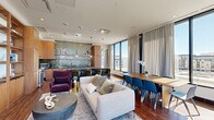 Located at 1901 Chestnut Place in Denver, Colorado, this single-floor open-plan space seamlessly integrates a kitchen, dining room, and living room into a cohesive, well-appointed environment. The layout maximizes functionality while maintaining a refined, contemporary aesthetic throughout.

The kitchen serves as a visual centerpiece, distinguished by a striking floor-to-ceiling green glazed tile backsplash that adds rich texture and color to the space. Warm-toned wood cabinetry pairs with a substantial dark marble island, underneath-lit for dramatic effect, and complemented by sage green upholstered barstools. Open floating shelves, pendant lighting, and stainless steel appliances complete the thoughtfully designed culinary area.

The dining zone features a natural wood table surrounded by modern blue chairs, positioned directly adjacent to expansive floor-to-ceiling windows that flood the space with abundant natural light and frame elevated views of the Denver skyline. Elegant floor-length drapery softens the glazed perimeter.

The living area offers a comfortable lounge arrangement anchored by a neutral sofa, plush purple accent chair, and a concrete round coffee table. A built-in wood shelving unit with integrated media storage lines one wall, grounding the space. Glass sliding doors open directly onto an outdoor terrace, extending the space and providing panoramic city views.