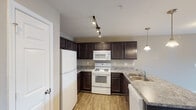This single-floor one-bedroom residence in Burlington, NC offers a thoughtfully designed layout that balances comfort and functionality. The kitchen is a standout feature, showcasing rich dark espresso cabinetry that contrasts beautifully with light granite-style countertops. White appliances — including a refrigerator, electric range, built-in microwave, and dishwasher — are complemented by a double-basin stainless steel sink and an extended breakfast bar peninsula. Track lighting overhead and elegant pendant lights add a polished, well-lit ambiance to the space. The open-concept living area is generously proportioned, finished with soft wall-to-wall carpeting in a neutral tone that enhances the room's warm, inviting feel. A ceiling fan with integrated lighting provides both comfort and illumination. Natural light pours in through a large window, while a set of French doors leads to a private balcony, offering a seamless indoor-outdoor connection. The bedroom is spacious and carpeted, featuring a ceiling fan and closet access. Throughout the residence, neutral warm-beige walls, white trim, and clean finishes create a cohesive, move-in-ready aesthetic suited to a variety of lifestyles.