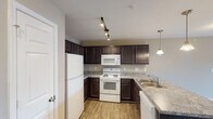 This single-floor one-bedroom residence in Burlington, NC offers a thoughtfully designed layout that balances comfort and functionality. The kitchen is a standout feature, showcasing rich dark espresso cabinetry that contrasts beautifully with light granite-style countertops. White appliances — including a refrigerator, electric range, built-in microwave, and dishwasher — are complemented by a double-basin stainless steel sink and an extended breakfast bar peninsula. Track lighting overhead and elegant pendant lights add a polished, well-lit ambiance to the space. The open-concept living area is generously proportioned, finished with soft wall-to-wall carpeting in a neutral tone that enhances the room's warm, inviting feel. A ceiling fan with integrated lighting provides both comfort and illumination. Natural light pours in through a large window, while a set of French doors leads to a private balcony, offering a seamless indoor-outdoor connection. The bedroom is spacious and carpeted, featuring a ceiling fan and closet access. Throughout the residence, neutral warm-beige walls, white trim, and clean finishes create a cohesive, move-in-ready aesthetic suited to a variety of lifestyles.