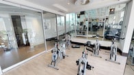 This single-floor facility offers a thoughtfully designed space that combines fitness and comfort in a clean, contemporary aesthetic. The fitness area is bright and open, featuring a dedicated cycling studio with multiple stationary bikes arranged on light hardwood-style flooring. Floor-to-ceiling glass partitions separate the cycling area from a fully equipped weight and strength training zone, which is lined with large wall mirrors that amplify the sense of space and allow for proper form monitoring. Ceiling fans provide airflow throughout the gym area, contributing to a comfortable workout environment. The bathroom is finished to a high standard, showcasing granite countertops with an undermount sink, soft-close cabinetry in a crisp white finish, and large-format tile flooring in a cool gray tone. A full-width mirror above the vanity is illuminated by modern lighting, enhancing the functionality and feel of the space. The tub and shower combination features classic white subway tile accented by a gray tile border, complemented by brushed nickel fixtures. Tasteful abstract artwork adds a refined touch throughout the bathroom, creating a polished and welcoming atmosphere that balances practicality with style.