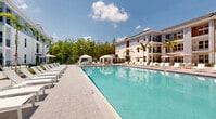 Located at 3240 Champion Ring Rd. in Fort Myers, Florida, this outdoor amenity space showcases a resort-inspired design perfectly suited to Southwest Florida living. At its centerpiece is a generously sized swimming pool with clear, turquoise water, surrounded by an expansive paver pool deck lined with white lounge chairs and shaded by elegant canopy cabanas — ideal for relaxing in the Florida sunshine.

The surrounding courtyard is thoughtfully landscaped with towering palm trees, tropical shrubs, and manicured garden beds set in white gravel, creating a lush, tropical ambiance throughout. Multiple outdoor seating areas with tables and umbrellas offer comfortable spaces for lounging and socializing, while iron-gated entryways provide secure access to the pool area.

The multi-story building enclosing the courtyard features a contemporary coastal architectural style, with a neutral color palette of white, beige, and slate blue, accented by black iron railings on private balconies that overlook the amenity space below. Large windows and glass doors connect interior common areas to the outdoor courtyard, enhancing the sense of openness. Mature trees line the perimeter beyond the fencing, providing natural privacy and a verdant backdrop that complements the community's polished, resort-like atmosphere.