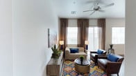This single-floor residence offers a comfortable and thoughtfully designed living space that blends modern style with warm, inviting touches. The living area serves as the heart of the home, showcasing a neutral color palette anchored by crisp white walls and complemented by rich, earthy tones. A ceiling fan adds both function and a subtle architectural detail overhead, while large windows dressed in layered brown drapes and roman shades flood the room with generous natural light and provide access to an outdoor area beyond a sliding glass door. The living room is furnished with a plush beige sofa and deep brown leather accent chairs, all centered around a bold, multicolored medallion-patterned area rug that adds a vibrant focal point to the space. A round ottoman coffee table with a decorative green plant sits at the center, tying the arrangement together. Along one wall, a low-profile wooden console displays potted succulents and greenery, adding a fresh, natural element. Framed wall art featuring a landscape scene completes the décor. The overall ambiance is warm, curated, and welcoming, making it an ideal space for both relaxing and entertaining.