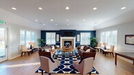 This single-floor space functions as a well-appointed community or gathering area, thoughtfully designed to balance comfort and functionality. The main lounge is the centerpiece, featuring a striking navy blue accent wall that frames a tiled fireplace with a white mantel and wall-mounted television above. Vaulted ceilings with recessed lighting lend the room an open, airy feel, while light hardwood flooring runs throughout, unifying the space. Multiple seating arrangements—including rich brown leather sofas, upholstered accent chairs, and a bold navy geometric area rug—create inviting conversation zones suited for both casual and social gatherings. Large windows fitted with white blinds line the walls, flooding the interior with natural light and offering views of the surrounding landscape.

A secondary seating nook near the entrance provides a quieter retreat, complete with upholstered chairs, a side table, and additional large windows. The entry area features decorative artwork and a dark wood console, adding a welcoming, curated aesthetic.

Adjacent to the main lounge, a kitchenette area offers dark cabinetry, granite-style countertops, appliances, and a pass-through counter, complemented by a small dining table nearby—making the space well-suited for events and everyday use.