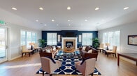 This single-floor space functions as a well-appointed community or gathering area, thoughtfully designed to balance comfort and functionality. The main lounge is the centerpiece, featuring a striking navy blue accent wall that frames a tiled fireplace with a white mantel and wall-mounted television above. Vaulted ceilings with recessed lighting lend the room an open, airy feel, while light hardwood flooring runs throughout, unifying the space. Multiple seating arrangements—including rich brown leather sofas, upholstered accent chairs, and a bold navy geometric area rug—create inviting conversation zones suited for both casual and social gatherings. Large windows fitted with white blinds line the walls, flooding the interior with natural light and offering views of the surrounding landscape.

A secondary seating nook near the entrance provides a quieter retreat, complete with upholstered chairs, a side table, and additional large windows. The entry area features decorative artwork and a dark wood console, adding a welcoming, curated aesthetic.

Adjacent to the main lounge, a kitchenette area offers dark cabinetry, granite-style countertops, appliances, and a pass-through counter, complemented by a small dining table nearby—making the space well-suited for events and everyday use.
