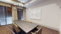 This single-floor space showcases a thoughtfully designed interior centered around a striking conference or dining area. The room features a large, dark matte-finish rectangular table surrounded by upholstered seating in warm golden and taupe tones, offering both comfort and style. Overhead, a sculptural multi-arm chandelier with exposed bulbs serves as a bold focal point, combining industrial and contemporary design elements to illuminate the space effectively.

Crown molding along the ceiling adds a refined architectural detail, lending a polished character to the room. The neutral palette of warm whites and greiges on the walls creates a calm, professional atmosphere, complemented by rich hardwood-style flooring that grounds the space with warmth and texture.

A large-scale abstract artwork adorns one wall, introducing artistic depth while maintaining the space's understated elegance. Natural light filters in through a window dressed with horizontal blinds and floor-length drape panels, allowing for flexible light control. The overall ambiance is sophisticated and contemporary, balancing functionality with refined aesthetic sensibility.