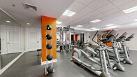 This fitness center at 25 Glenbrook Rd in Stamford, Connecticut, offers a well-appointed, single-floor exercise space designed to support a full range of workout routines. The room features durable rubber flooring throughout, providing a safe and functional surface for both cardio and strength training activities. A dropped ceiling with recessed panel lighting ensures consistent, even illumination across the entire space.

The cardio area is generously equipped with multiple treadmills, elliptical trainers, and stationary bikes, arranged to allow comfortable spacing between machines. A wall-mounted flat-screen television is positioned for easy viewing during workouts. Large glass doors and windows along one wall allow natural light to filter in, creating an inviting and energizing atmosphere.

The strength training section includes a cable machine system, adjustable weight bench, and a full dumbbell rack, catering to users of varying fitness levels. Bold orange accent walls add a vibrant, motivating energy to the otherwise clean white interior. Floor-to-ceiling mirrors in the strength area enhance the sense of space and allow users to monitor their form. The room is accessed through elegant French doors, adding a refined architectural detail to this practical and fully functional fitness facility.