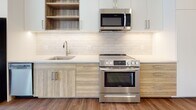 Located at 3009 W Leigh St in Richmond, Virginia, this single-floor unit presents a clean, modern aesthetic with thoughtfully designed living spaces. The layout, as seen in the floor plan, organizes the rooms efficiently, connecting the main living areas with ease.

The kitchen is a standout feature, showcasing stainless steel appliances including a full-size refrigerator, electric range, and over-the-range microwave. Light wood cabinetry is paired with white upper cabinets and open shelving, complemented by a white subway tile backsplash and light-toned countertops. Wood-look plank flooring grounds the space and extends into the adjoining living area, adding warmth to the contemporary design.

The bedroom features neutral wall-to-wall carpeting and a ceiling fan with integrated lighting, creating a comfortable, quiet retreat. The bathroom is finished with large-format white tile flooring, a tiled soaking tub and shower combination with a built-in niche, a wood-toned vanity with a clean white countertop, and a window that allows natural light to filter in. Throughout the unit, white walls and consistent modern finishes contribute to a bright, cohesive atmosphere.