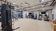 This modern fitness center offers a generously sized, open-plan workout space designed to accommodate a full range of exercise activities. The room features durable, light-toned rubberized flooring that provides both comfort and functionality underfoot, complemented by smooth gray walls and clean white trim throughout.

A striking design feature is the series of large circular ring pendant light fixtures suspended from the ceiling, adding an industrial-chic aesthetic to the space. Exposed ceiling infrastructure, recessed lighting, and track lighting work together to ensure the area is well-illuminated. A bold textured accent wall in deep blue-gray tones adds visual depth and a contemporary feel to one side of the room.

Large wood-framed mirrors line a significant portion of the walls, enhancing the sense of openness and allowing users to monitor their form during workouts. Mounted flat-screen televisions are positioned for easy viewing throughout the space.

The fitness center is equipped with a comprehensive selection of equipment, including treadmills, stationary spin bikes, a stair climber, an elliptical, a cable machine, a power rack, and adjustable benches. A dedicated storage shelving unit provides organized space for accessories and supplies. A keyed entry door with a glass window panel ensures secure, controlled access to the facility.