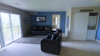 This single-floor space centers around a well-appointed living room that makes a strong visual impression through its use of a deep blue accent wall, which anchors the room and adds a sense of depth and character. The living area is furnished with a matching set of dark leather sofas and chairs arranged on a textured area rug over neutral carpeting, creating a cohesive and comfortable seating arrangement. Framed artwork decorates the walls, contributing to a polished, curated feel throughout the space. A ceiling vent is visible overhead, indicating a central HVAC system. The room flows naturally into a hallway that leads to additional areas of the home, hinting at a connected and functional layout. Neutral-toned walls in the adjoining spaces complement the bolder styling of the main living area, offering visual balance. A closet with a bi-fold door is visible along one wall, providing convenient storage. The overall ambiance is contemporary and inviting, with a clear emphasis on comfort and style through the deliberate use of color contrast and coordinated furnishings.