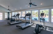 Located at 7227 N Mesa St in El Paso, Texas, this single-story community facility offers a thoughtfully designed space that combines fitness and social amenities. The fitness area is the centerpiece of the space, featuring vaulted ceilings that lend an open, airy atmosphere. It is well-lit by natural light streaming through multiple large windows overlooking the outdoor surroundings. The gym is equipped with multiple treadmills, a stationary bike, and a cable weight machine, all set atop durable carpeted flooring. A ceiling fan ensures comfortable airflow during workouts. A striking exposed brick accent wall adds architectural character, anchoring a workstation area outfitted with a large flat-screen TV, a desk, and ergonomic office chairs — ideal for business or study use. Pendant lighting above this area complements the rustic charm of the brickwork. A separate entry zone with tile flooring provides a transition space near the main entrance door, which features a decorative oval glass insert. The kitchenette area includes white cabinetry, granite-style countertops, stainless steel appliances including a refrigerator, and a breakfast bar with leather-upholstered bar stools, making it a practical space for gatherings or quick refreshments.