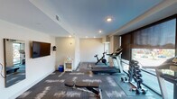 Located at 6311 E Westfield Blvd in the Broad Ripple area of Indianapolis, Indiana, this fitness center is a thoughtfully designed, single-floor exercise space built to support a well-rounded workout routine. The room features a clean, modern aesthetic with neutral-toned walls, recessed lighting, and patterned carpet tiles in shades of gray and brown that add visual texture underfoot.

The space is equipped with multiple cardio machines, including treadmills and a Peloton stationary bike, positioned near large floor-to-ceiling windows that allow abundant natural light and provide views of the surrounding neighborhood. A dumbbell rack with a selection of free weights lines one wall, offering options for strength training. Additional accessories such as resistance bands and ab benches are available for varied exercise routines.

A wall-mounted flat-screen television is positioned prominently for viewing during workouts, accompanied by a wall mirror that enhances the sense of openness. A small supply station with sanitation essentials is conveniently located within the room. The space is serviced by a dedicated HVAC system with ceiling vents, ensuring a comfortable environment year-round. A single entry door with a window provides secure, straightforward access. Overall, the fitness center delivers a functional and inviting environment for daily exercise.